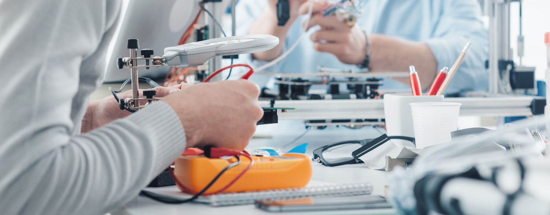 Engineering students working in a lab, a student is using a voltage and current tester, another student in the background is using a 3D printer